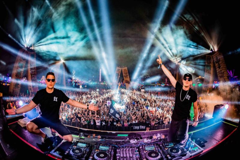Two DJs on a festival stage at night, arms raised, with a large crowd and light beams behind them