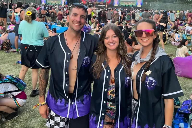 Three smiling customers wearing matching black baseball jerseys with purple forest artwork at a festival.