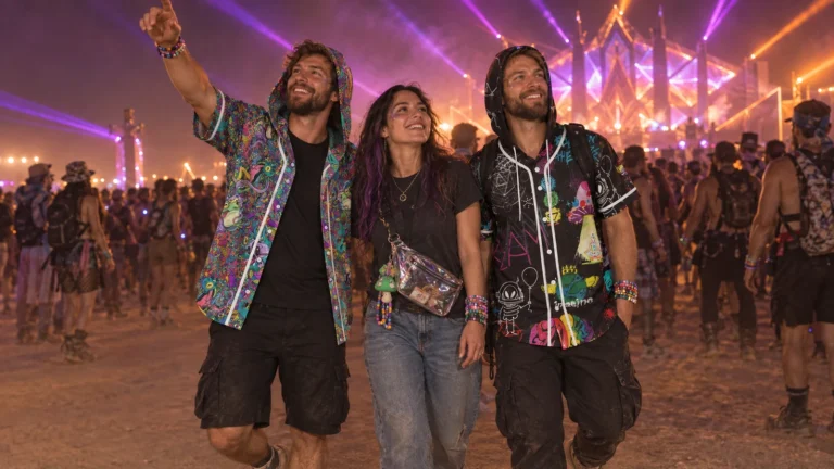 Three friends at a desert night festival, two of them in a psychedelic teal and purple hooded baseball jersey and a black hooded jersey with neon doodles, with stage lights behind them