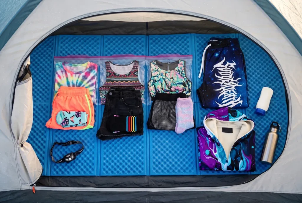 Festival camping checklist packing: three day outfits on a camping pad, each top in a clear zip bag with its shorts and socks below it, beside black galaxy-print rave joggers and a folded purple marbled rave cloak