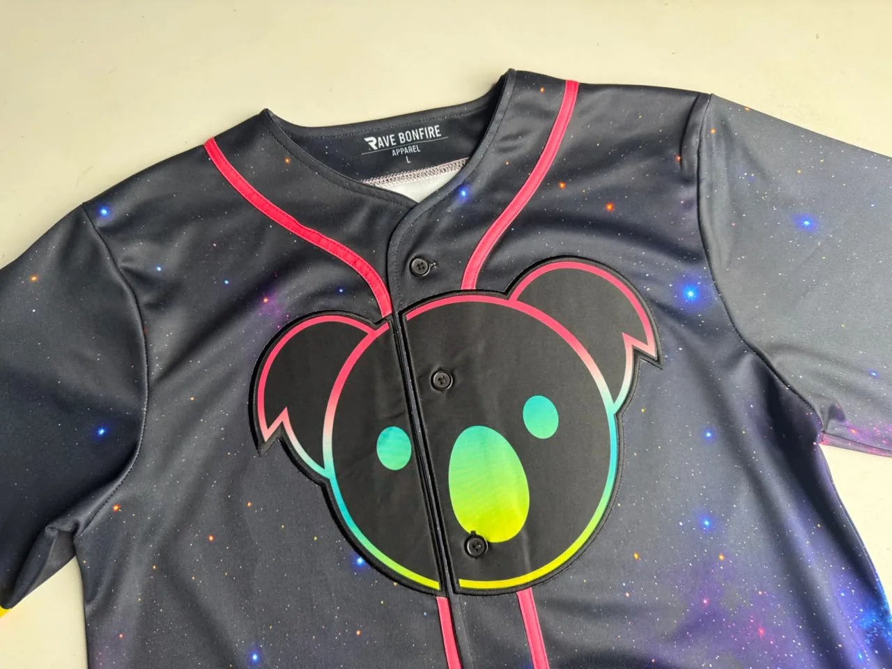 Front of a black galaxy-print baseball jersey with a large koala-head chest emblem across the button placket and hot-pink piping