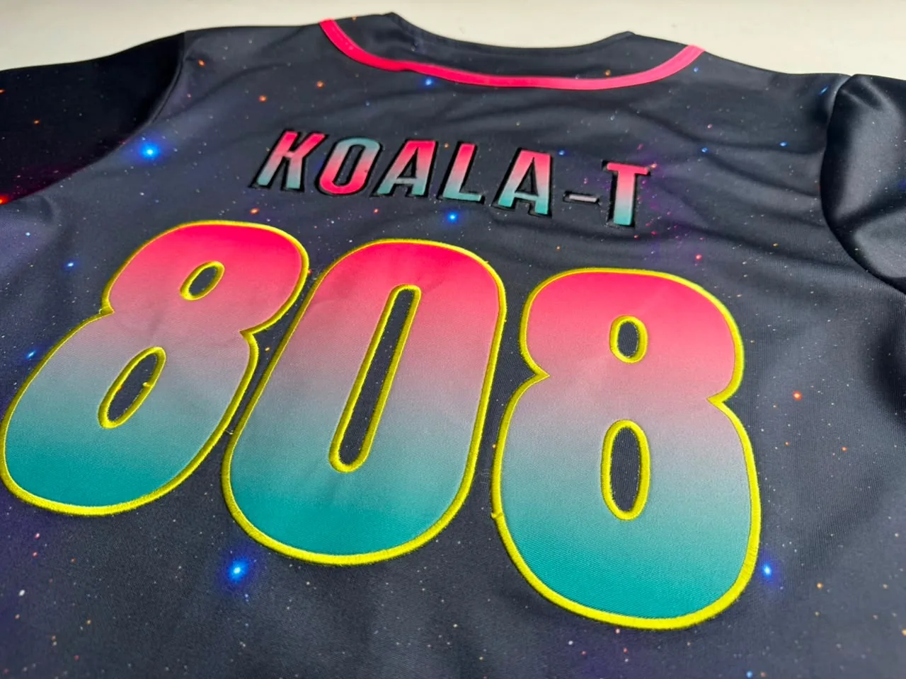 Close-up of a jersey back showing how rave names read: KOALA-T in pink-to-teal gradient letters over a large printed 808 with a raised yellow embroidered outline