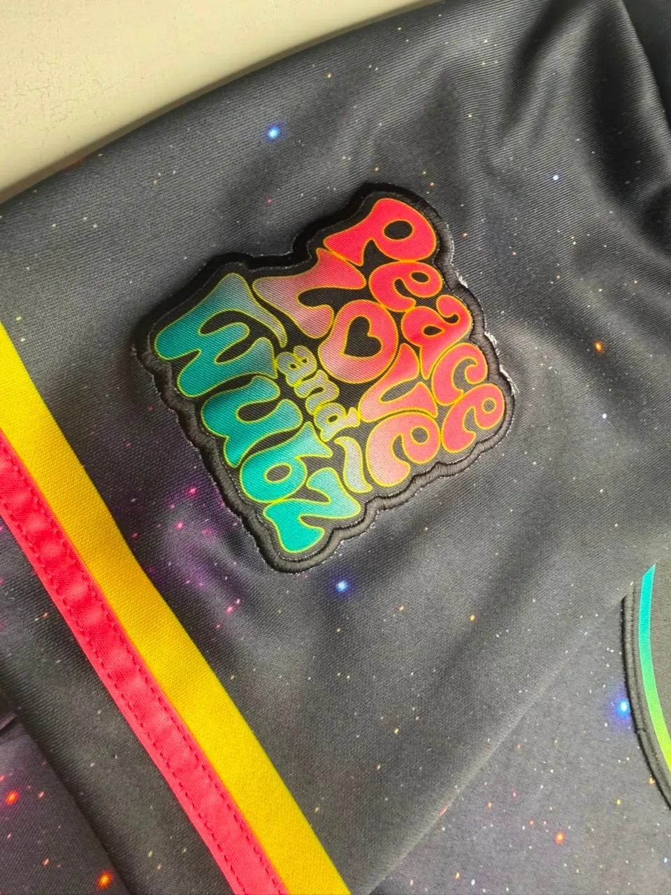 Sleeve of a galaxy-print jersey with a Peace Love and Wubz patch in pink-to-teal lettering and a stitched black edge above yellow and pink cuff stripes